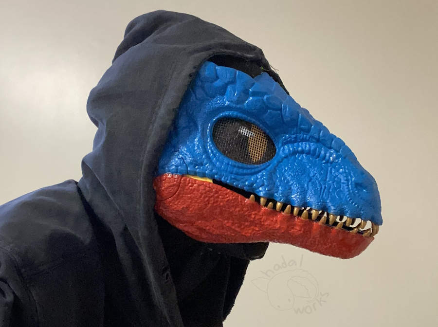 A plastic dinosaur mask with a custom paint job. The top is solid blue, and the bottom lower jaw is solid red. The teeth are shiny gold, and the membrane inside the mouth is yellow. Its mesh eyes are solid black with a shiny gold slit pupil and white eye gleam painted on.