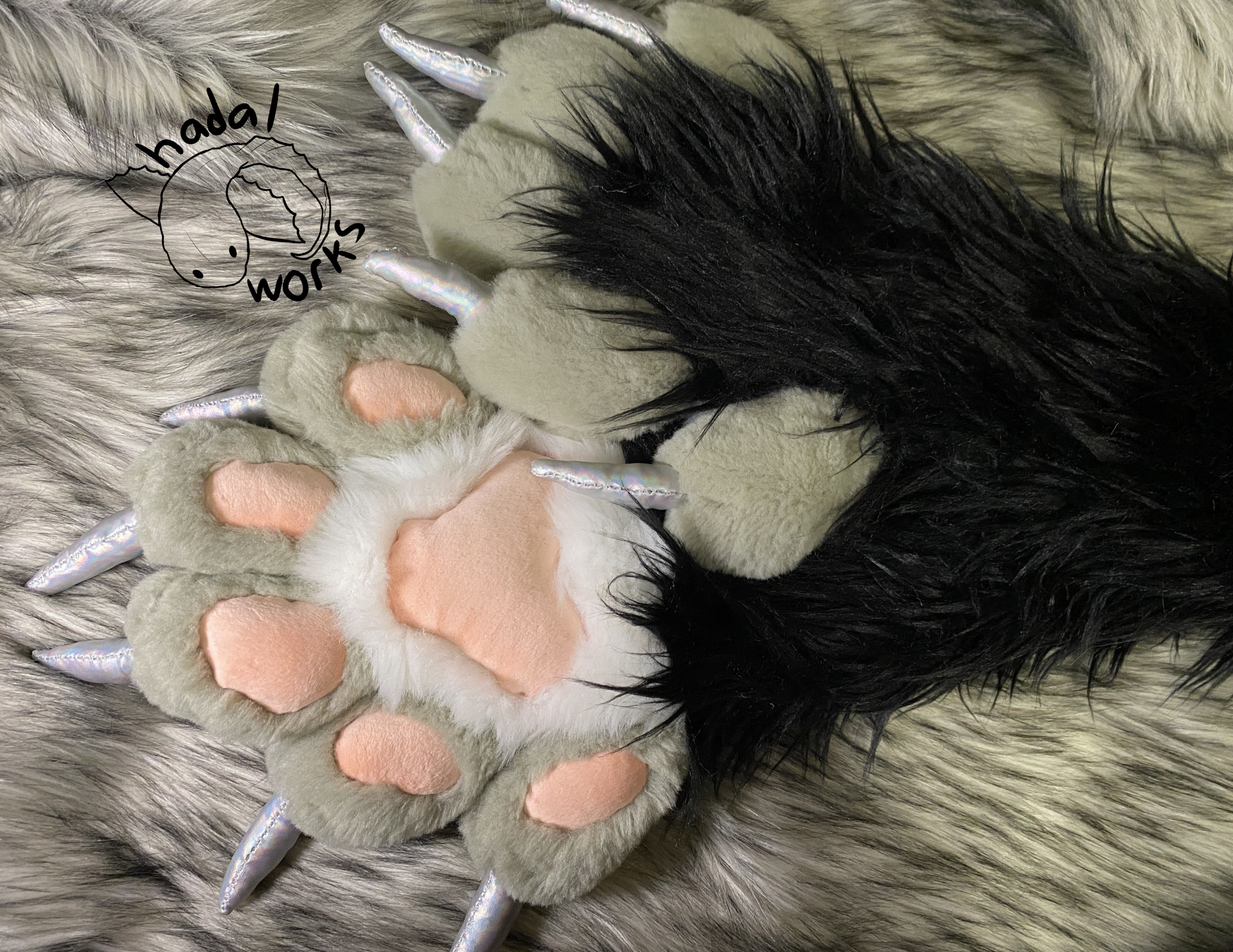 A pair of fursuit paws laying atop one another. The arm of the paws is black, with the fingers being gray. The palms are white. They have soft light pink minky pawpads. The claws are a holographic silver fabric. They lay on a long ticked gray fabric background.